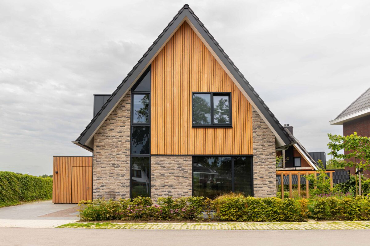 Vooraanzicht van de moderne schuurwoning met een combinatie van natuursteen en houten gevelbekleding, zwarte aluminium kozijnen en een strak afgewerkt dak met zonnepanelen.