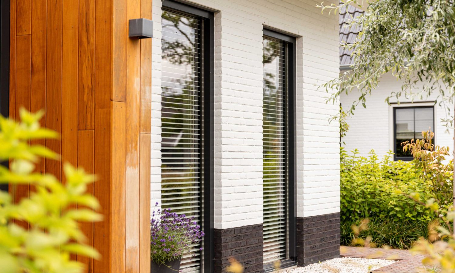 Detail van de gevel met hoekraam, wit gekeimde bakstenen en donkere plintsteen. Strakke afwerking met kozijnen in RAL 9021 en een grindkoffer voor een onderhoudsvriendelijke uitstraling.