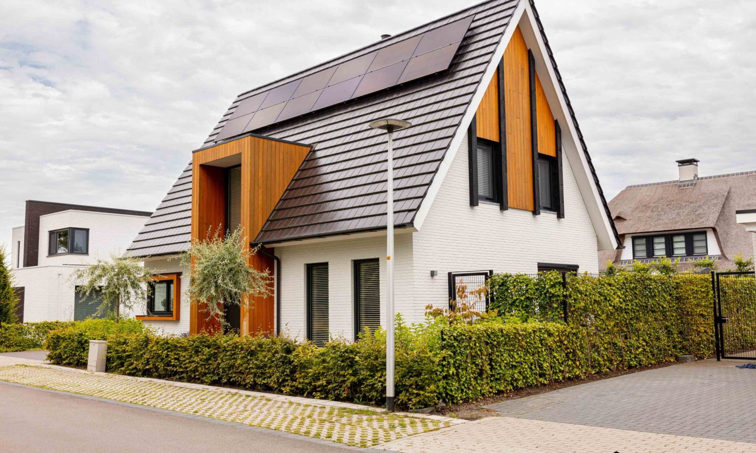 Eigentijdse vrijstaande woning in Hengelo, met wit gekeimde gevels, donkere plintsteen en risaliet met natuurlijke houtkleur. Vlakke keramische dakpan en aluminium bakgoten in RAL 9021 voor een strakke afwerking