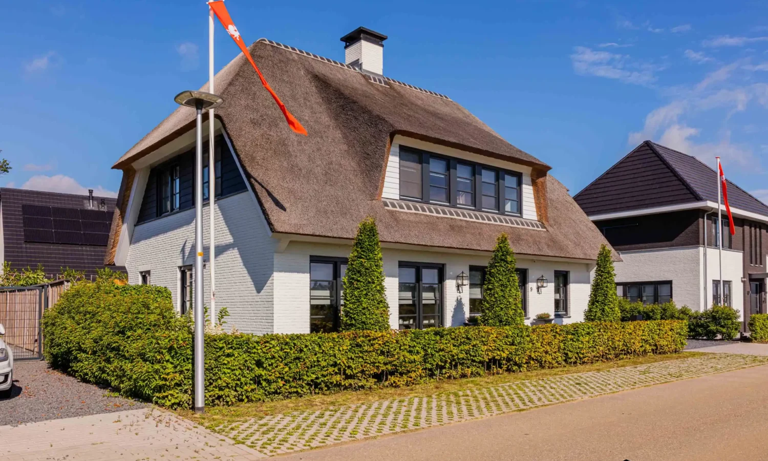 Buitenaanzicht van de woning met rieten dak en witte gevels, omringd door groene haag en bestrating