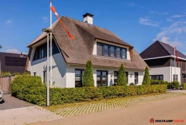 Buitenaanzicht van de woning met rieten dak en witte gevels, omringd door groene haag en bestrating