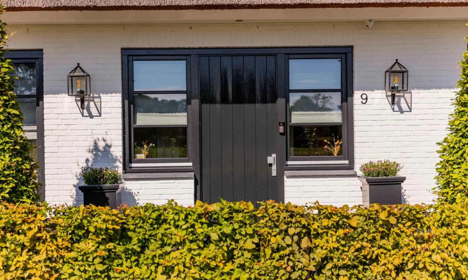 Moderne voordeur in zwarte tinten met karakteristieke wandlampen en hagen.